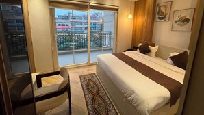 A hotel room with a large bed, a desk, and a view of the city through the balcony doors. - Hotel alvish (New Delhi)