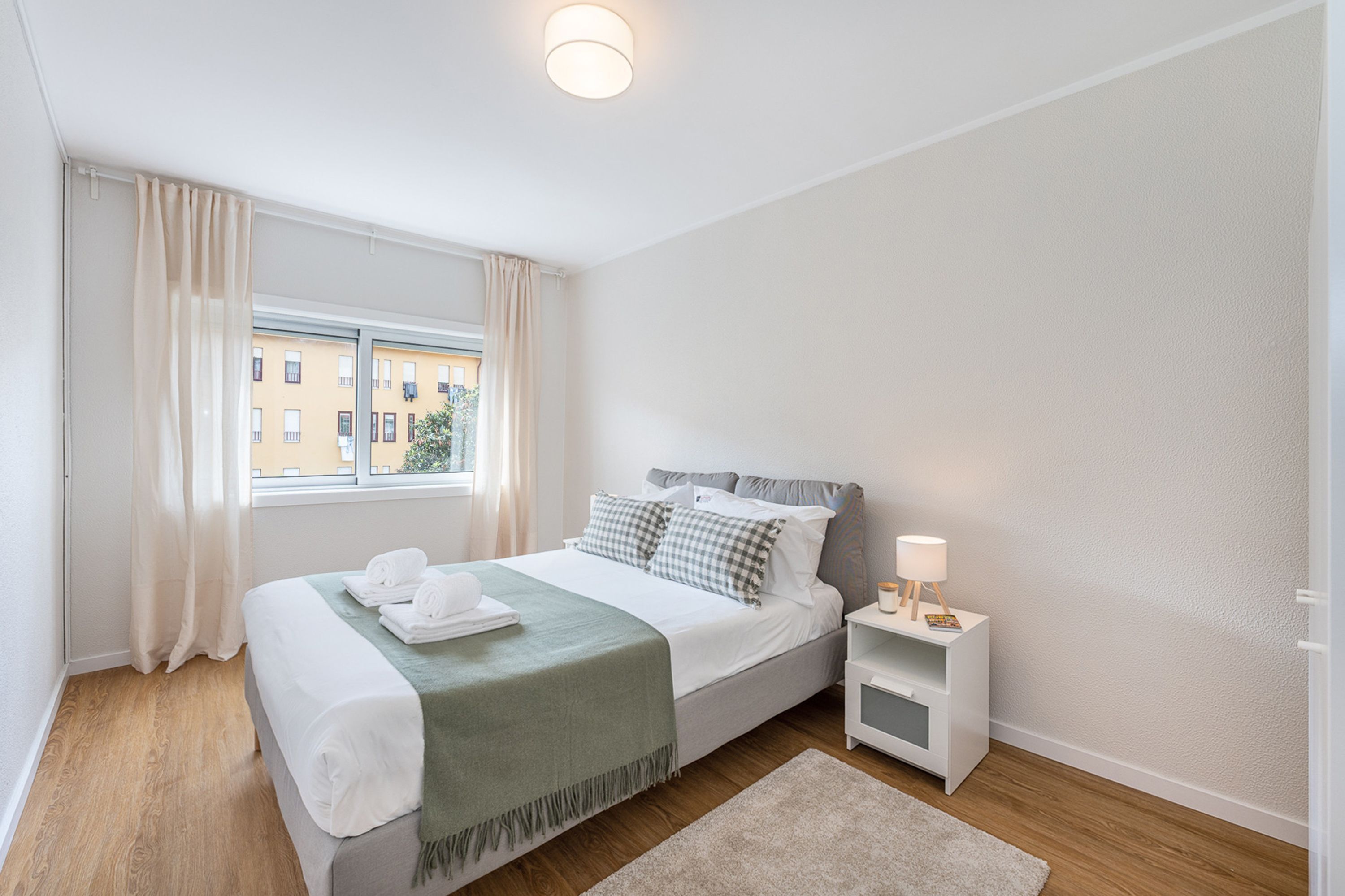 Una camera d'albergo con un letto, un comodino con una lampada, una finestra con tende e vista sugli edifici.
