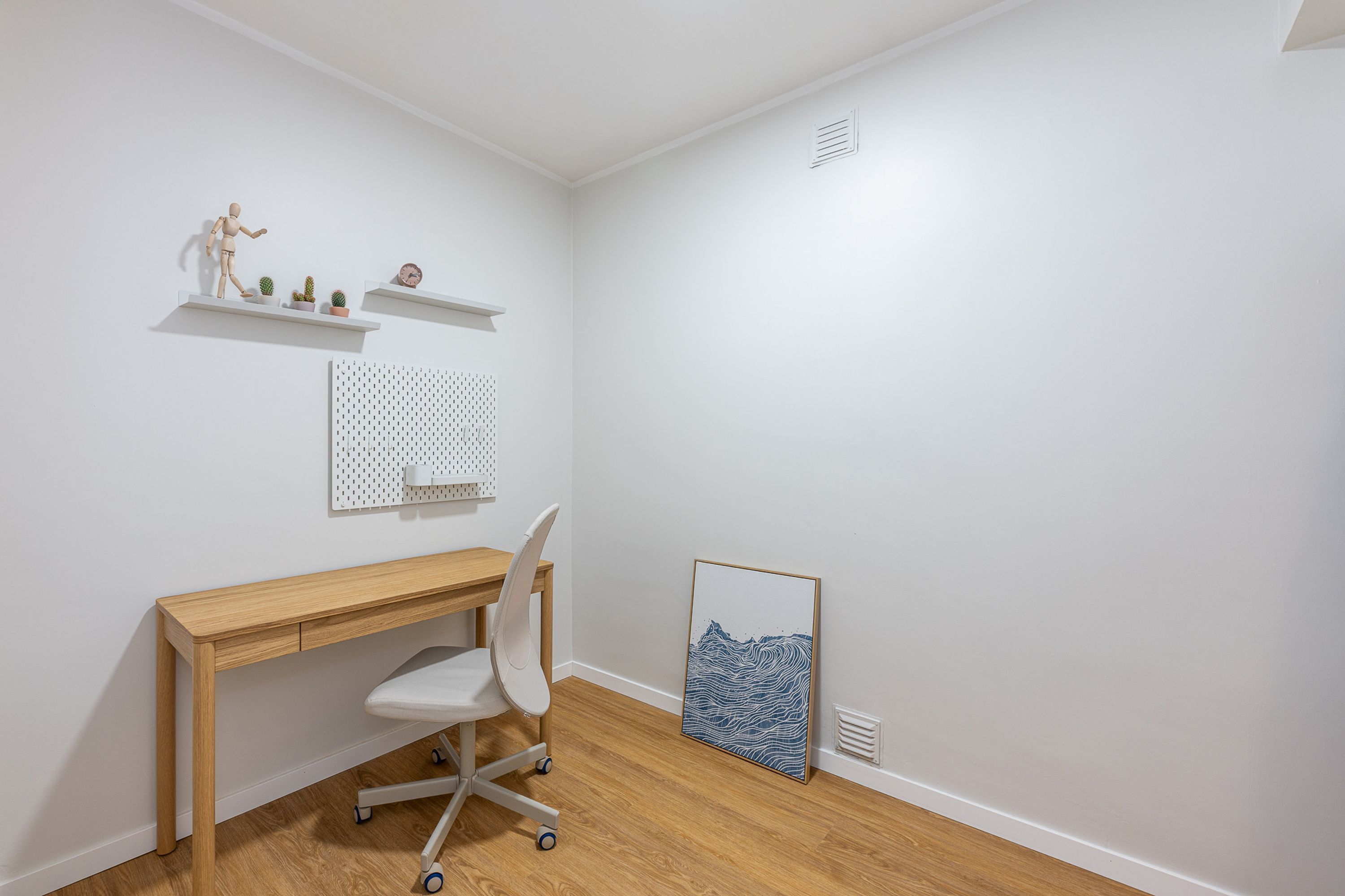 Een kamer met een houten bureau, een witte stoel, een prikbord en een lijst met een afbeelding.