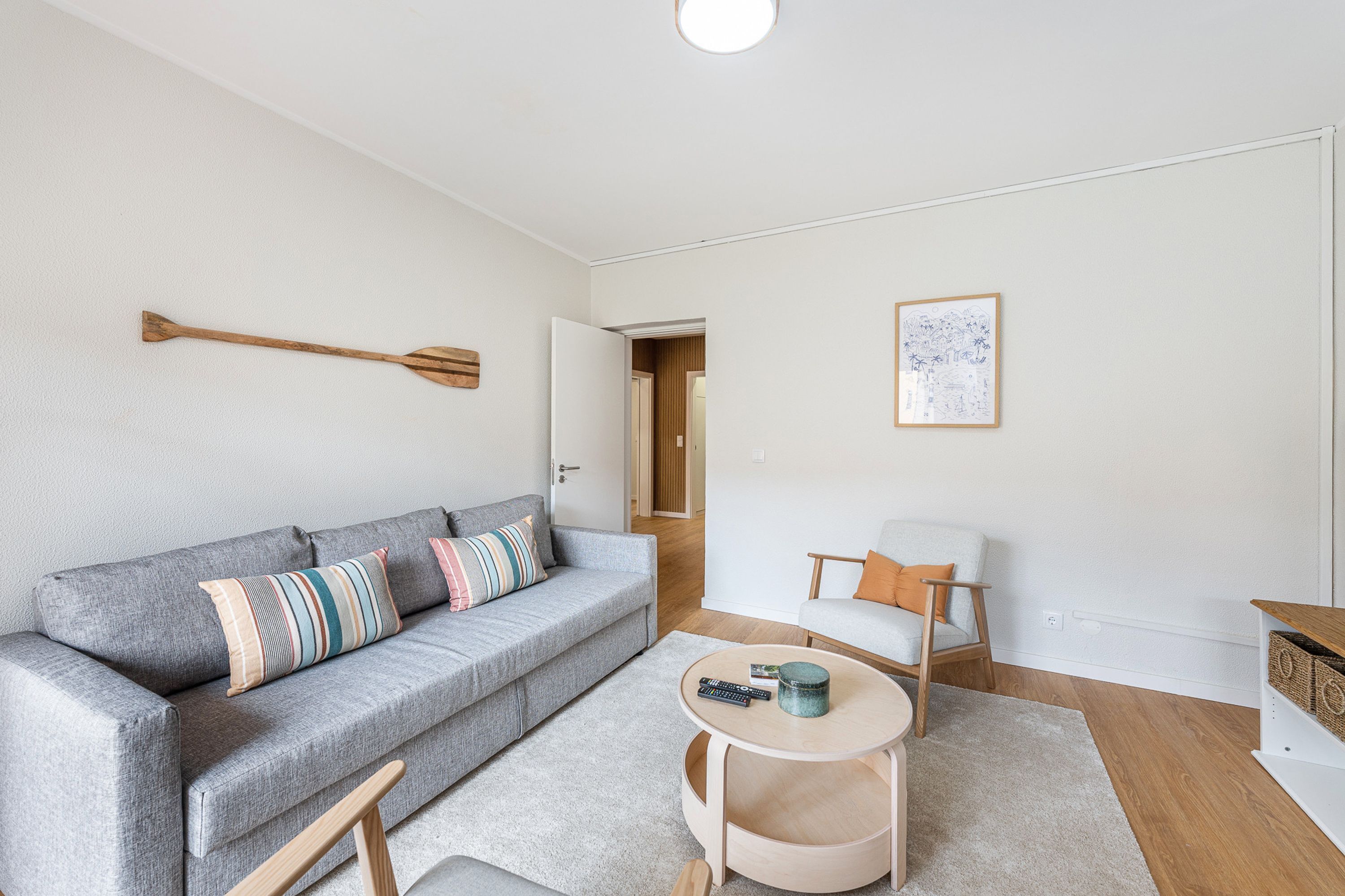 Een moderne woonkamer met een grijze bank, een klein rond bijzettafeltje, een houten nachtkastje en een lijst met een foto aan de muur.