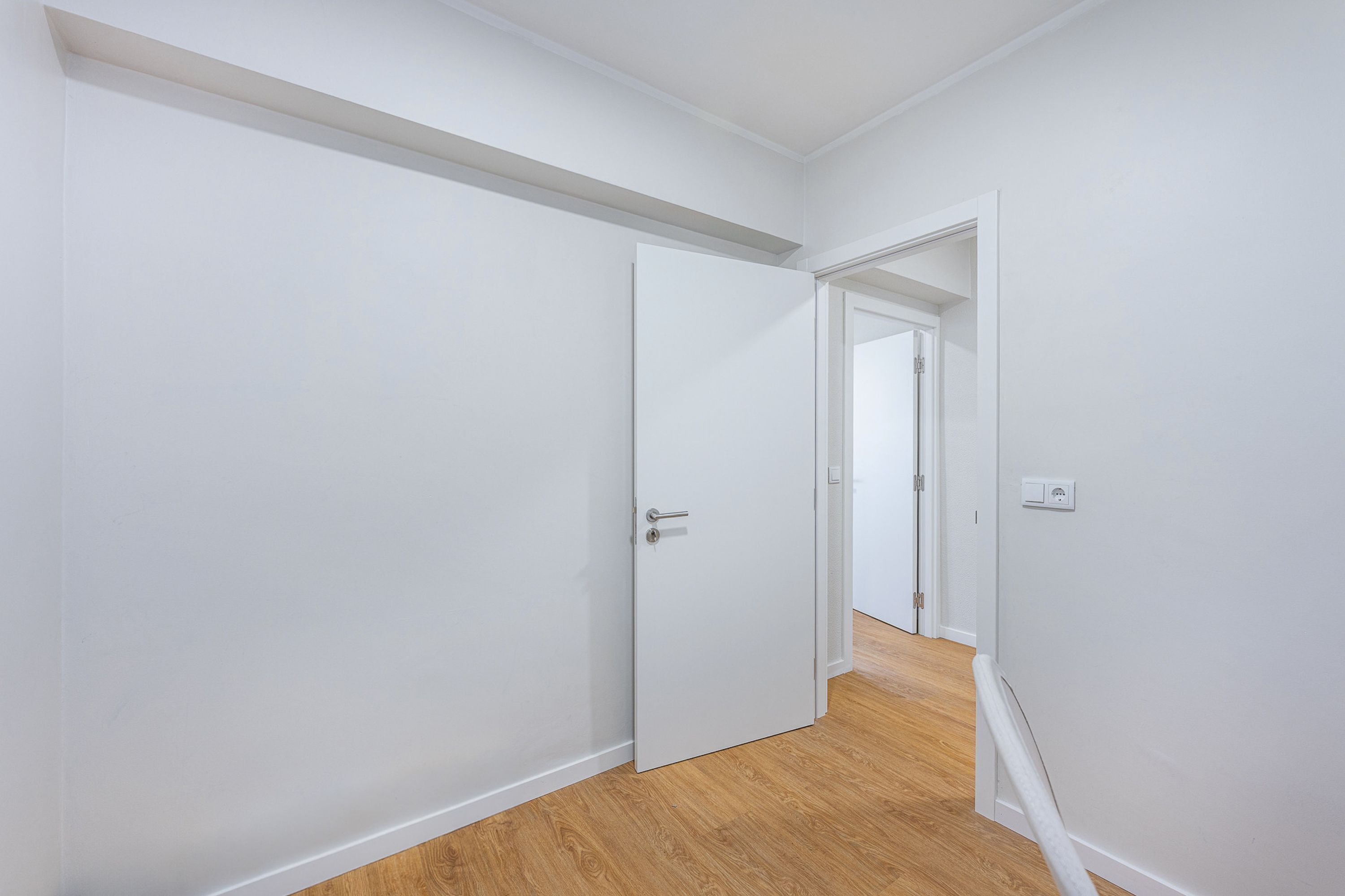A hallway with white walls and wooden flooring, leading to a room with a door.