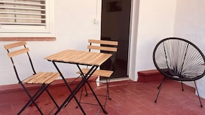 Eine Terrasse mit Holztisch und -stühlen, einem schwarzen Flechtstuhl und einem Fenster mit Jalousien.