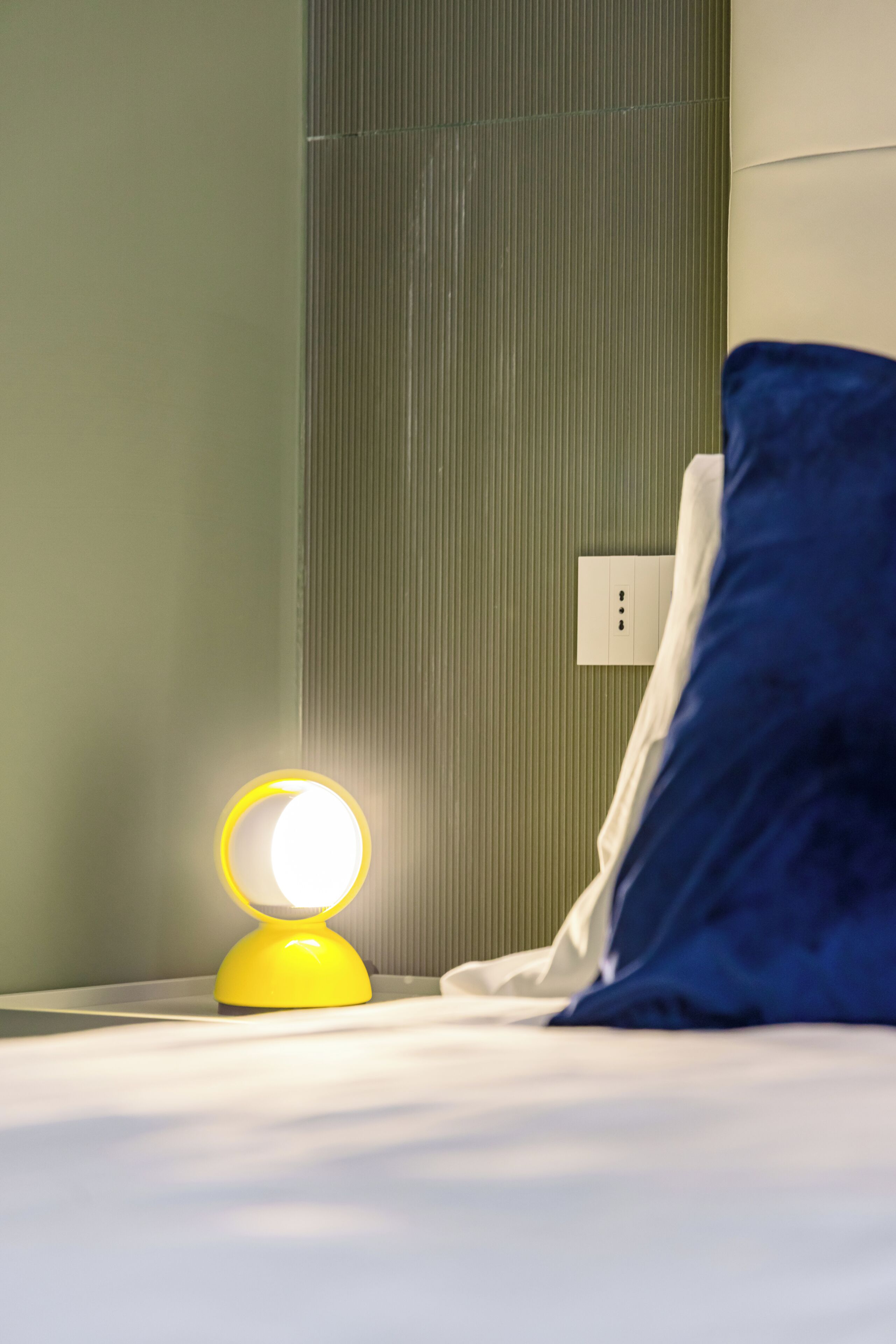 Un lit avec un drap blanc et un oreiller bleu, une lampe de chevet jaune et un mur orné de lignes verticales.