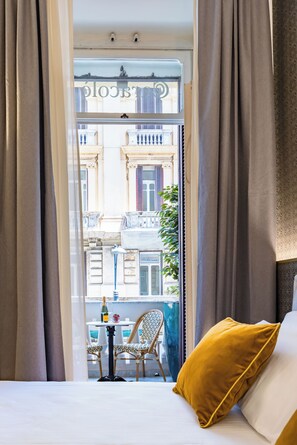 Habitación de hotel con cama, una mesita con una botella y vasos, una silla y vista a la calle por una ventana.