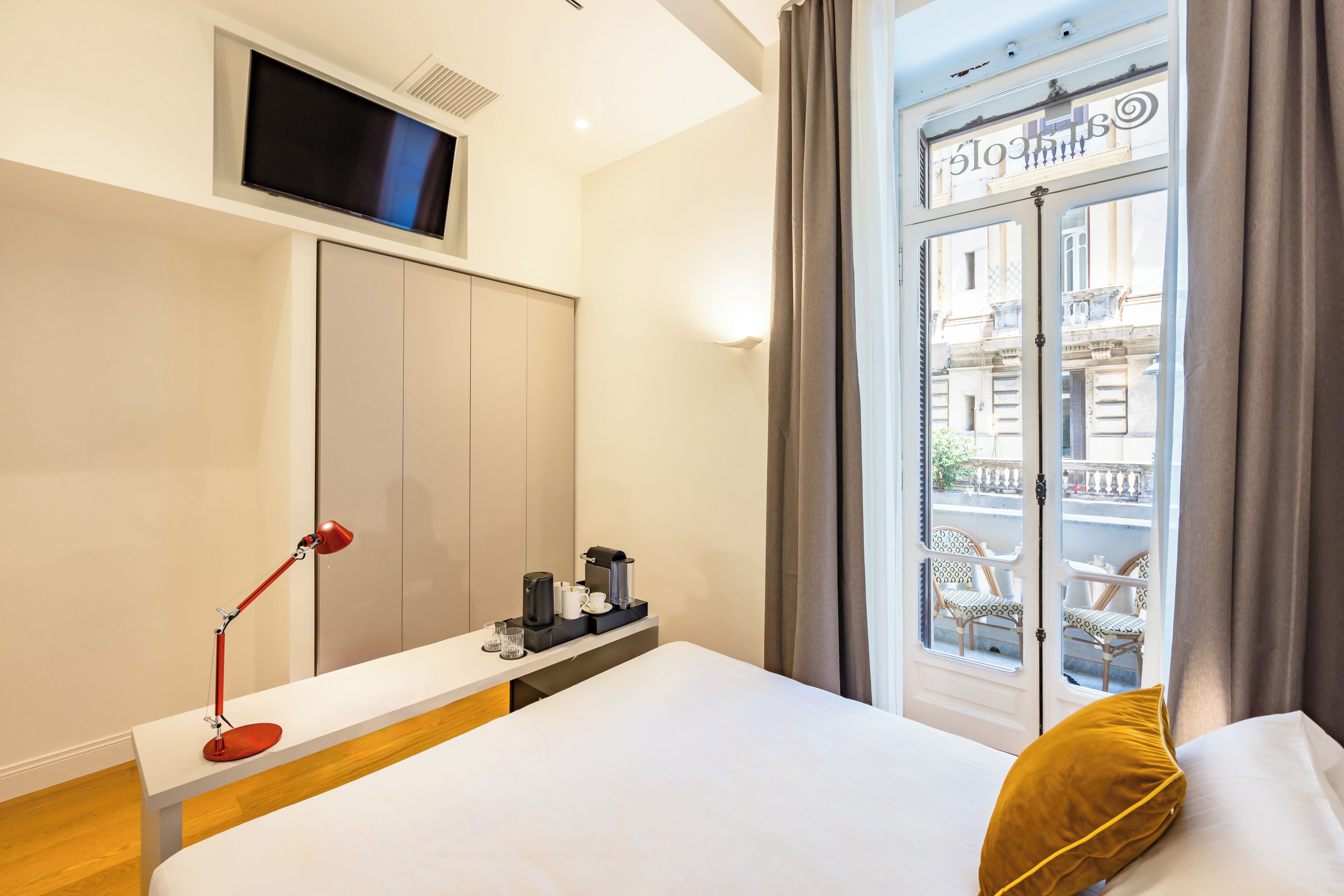 A hotel room with a bed, a desk with a lamp, a coffee maker, and a TV mounted on the wall.