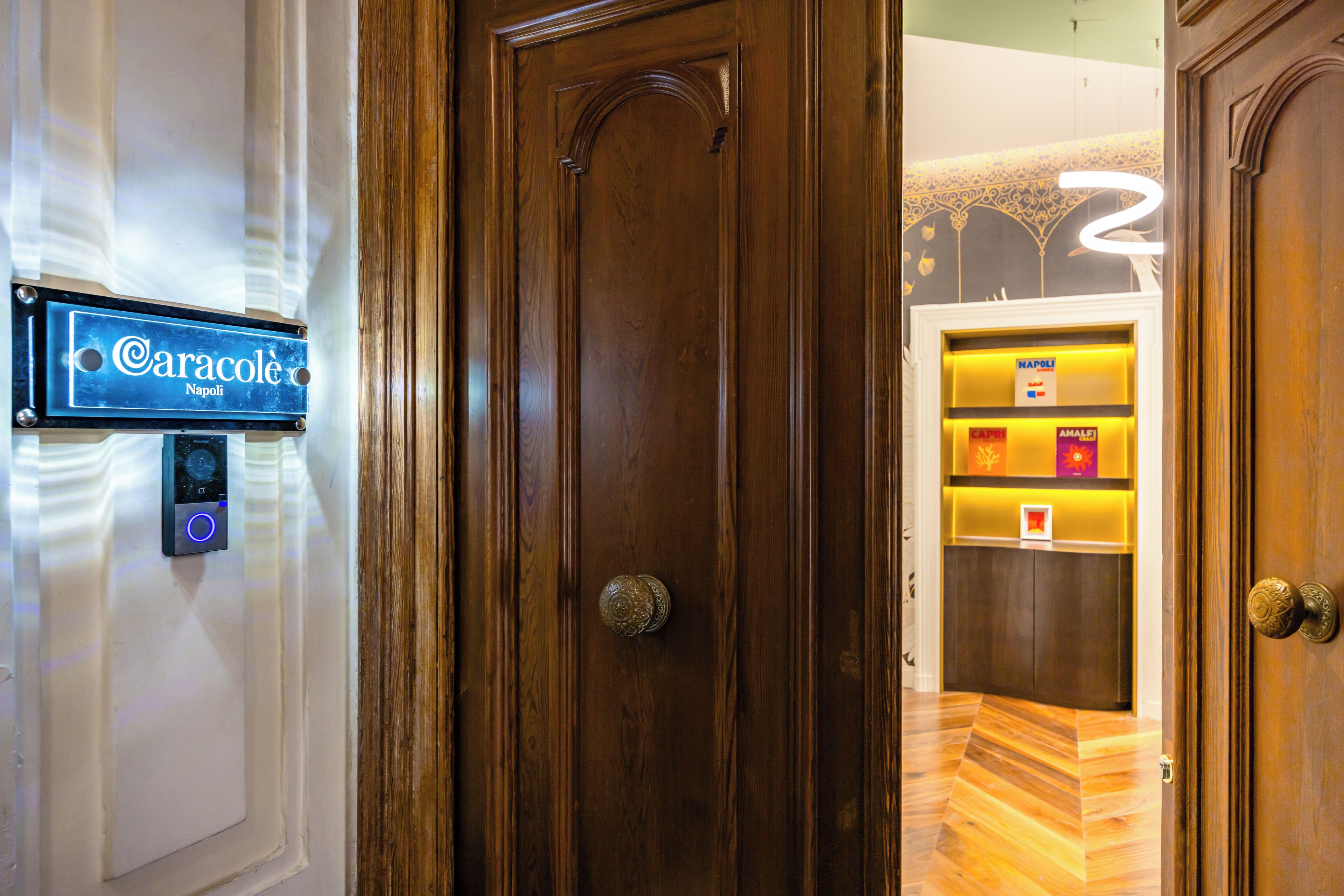 A hotel room entrance with a wooden door, a digital sign indicating 'Caracolé Napoli,' and a reception area with a counter and decorative elements.