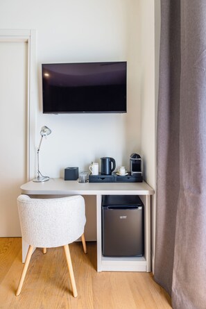 Habitación de hotel compacta con televisor de pantalla plana montado en la pared, una pequeña mesa de escritorio con lámpara, cafetera y lavavajillas pequeño.
