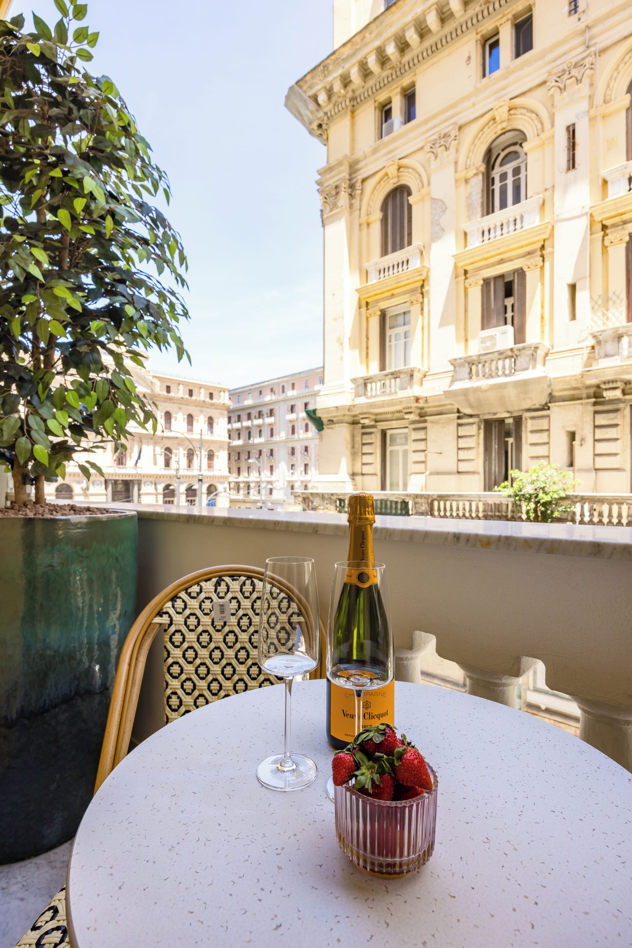 Une table avec une bouteille de champagne, un verre et un bol de fraises, le tout placé sur un balcon donnant sur un bâtiment historique.