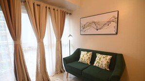 A living room with a green sofa, beige curtains, and a wall-mounted abstract painting. - Kuya Awens Staycation Unit 15K (Parañaque)