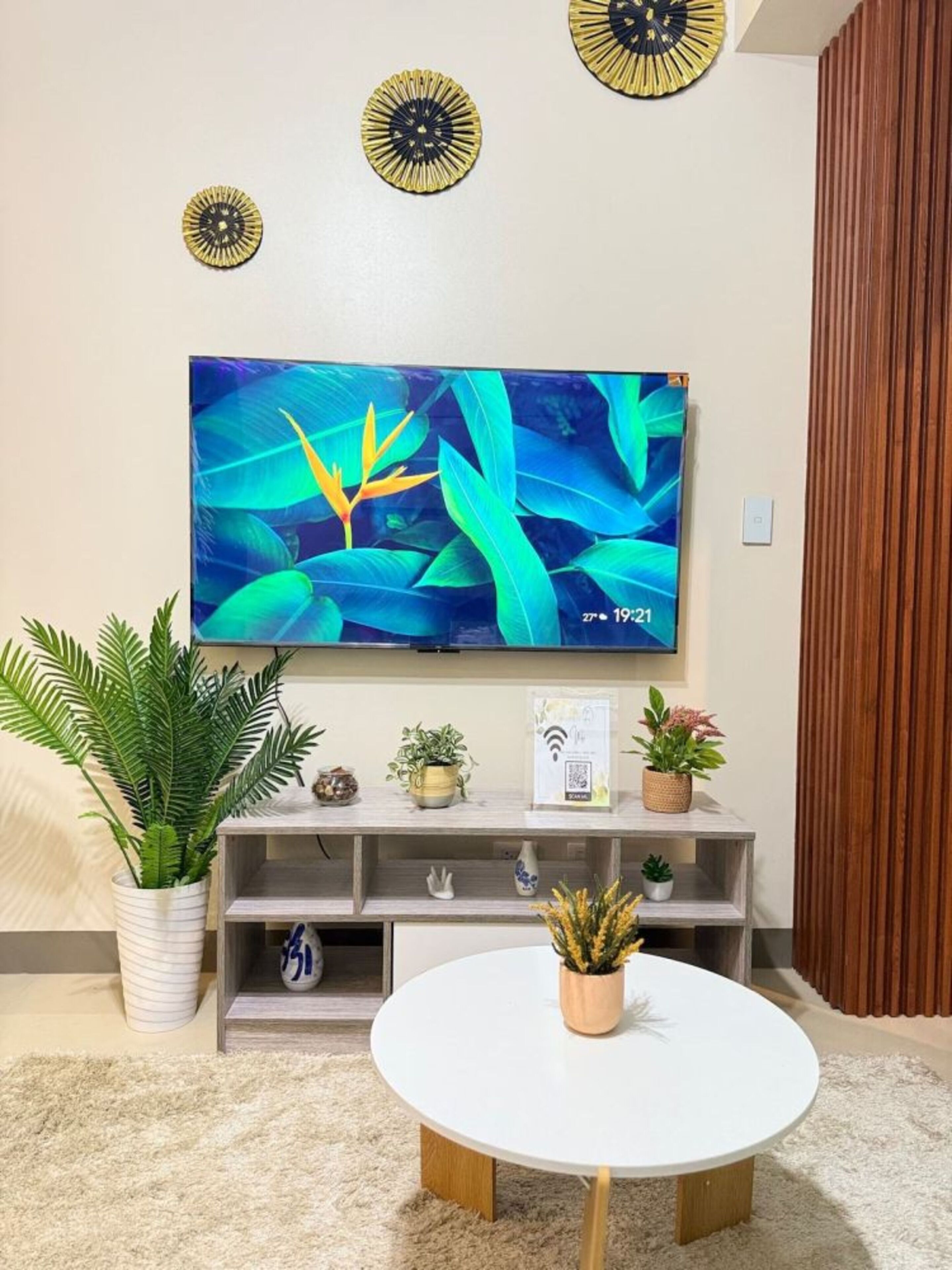 Una sala de estar con una televisión de pantalla plana montada en la pared, una mesa redonda blanca y una variedad de plantas en macetas sobre una consola de madera.