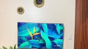 A living room with a flat-screen TV mounted on the wall, a round white table, and a variety of potted plants on a wooden console table. - Kuya Awens Staycation Unit 15K (Parañaque)