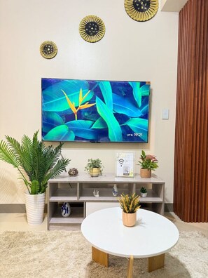 A living room with a flat-screen TV mounted on the wall, a round white table, and a variety of potted plants on a wooden console table. - Kuya Awens Staycation Unit 15K (Parañaque)