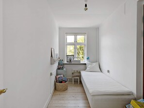Ein schmales Zimmer mit einem Einzelbett, einem kleinen Tisch, einem Stuhl und einem Fenster mit Blick ins Grüne.