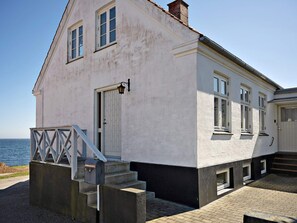 Ein zweistöckiges Gebäude mit weißer Fassade, eine Treppe zum Eingang und im Hintergrund ein Meerblick.