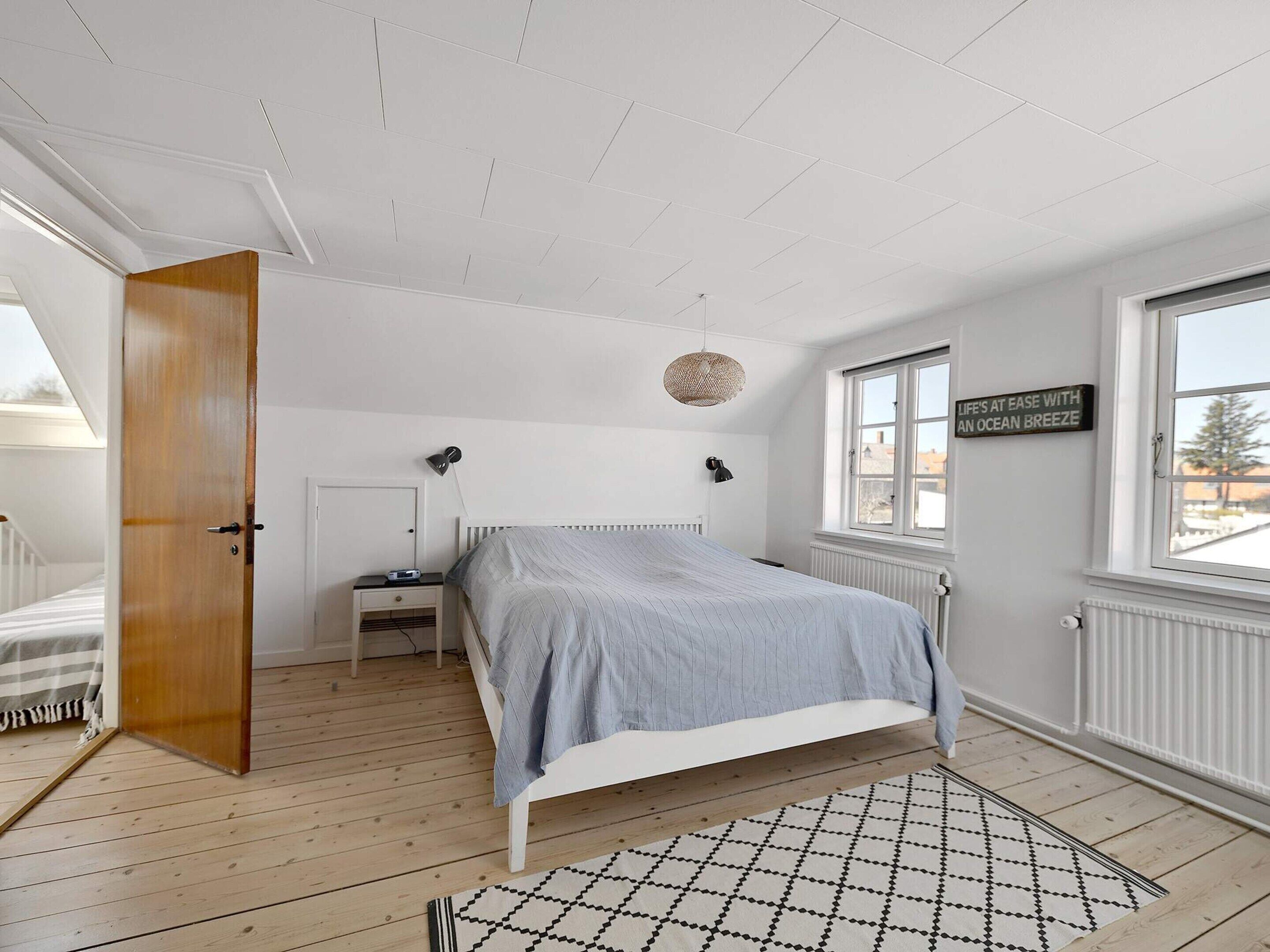 A bedroom with a bed, bedside tables, a hanging light fixture, a window with a view, and a door leading to another room.
