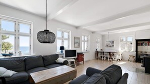 A spacious living room with a large sofa, a wooden coffee table, and a dining area with a table and chairs. - 5 Star Holiday Home in Svaneke (Svaneke)