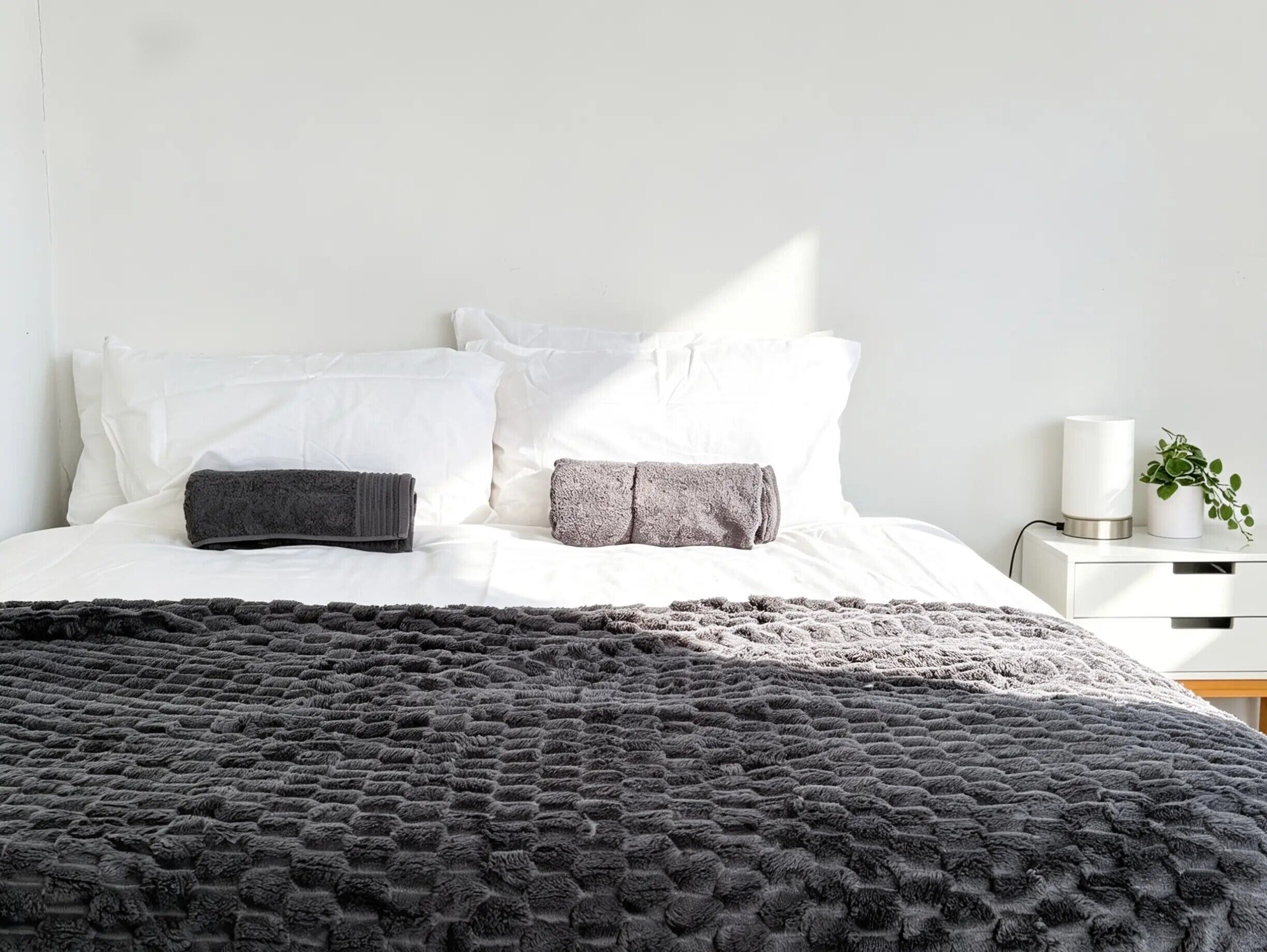 A bed with a textured blanket, white pillows, and a bedside table with a lamp and a plant.