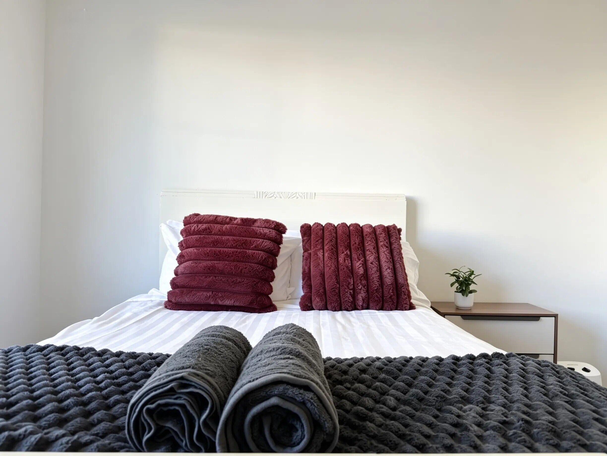 A neatly made bed with a white headboard, two rolled-up dark grey blankets, and two folded red towels.
