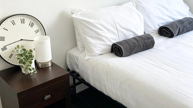 A neatly made bed with white linens and two rolled gray towels, a white wall clock, and a small plant on a bedside table. - Spacious Cosy Family & Pet-Friendly Home with Large Backyard (Dubbo)