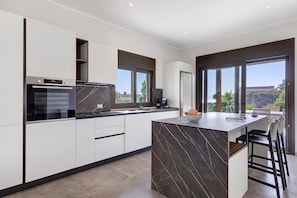 A modern kitchen with a marble island, built-in appliances, and large windows offering a view of the outdoors.
