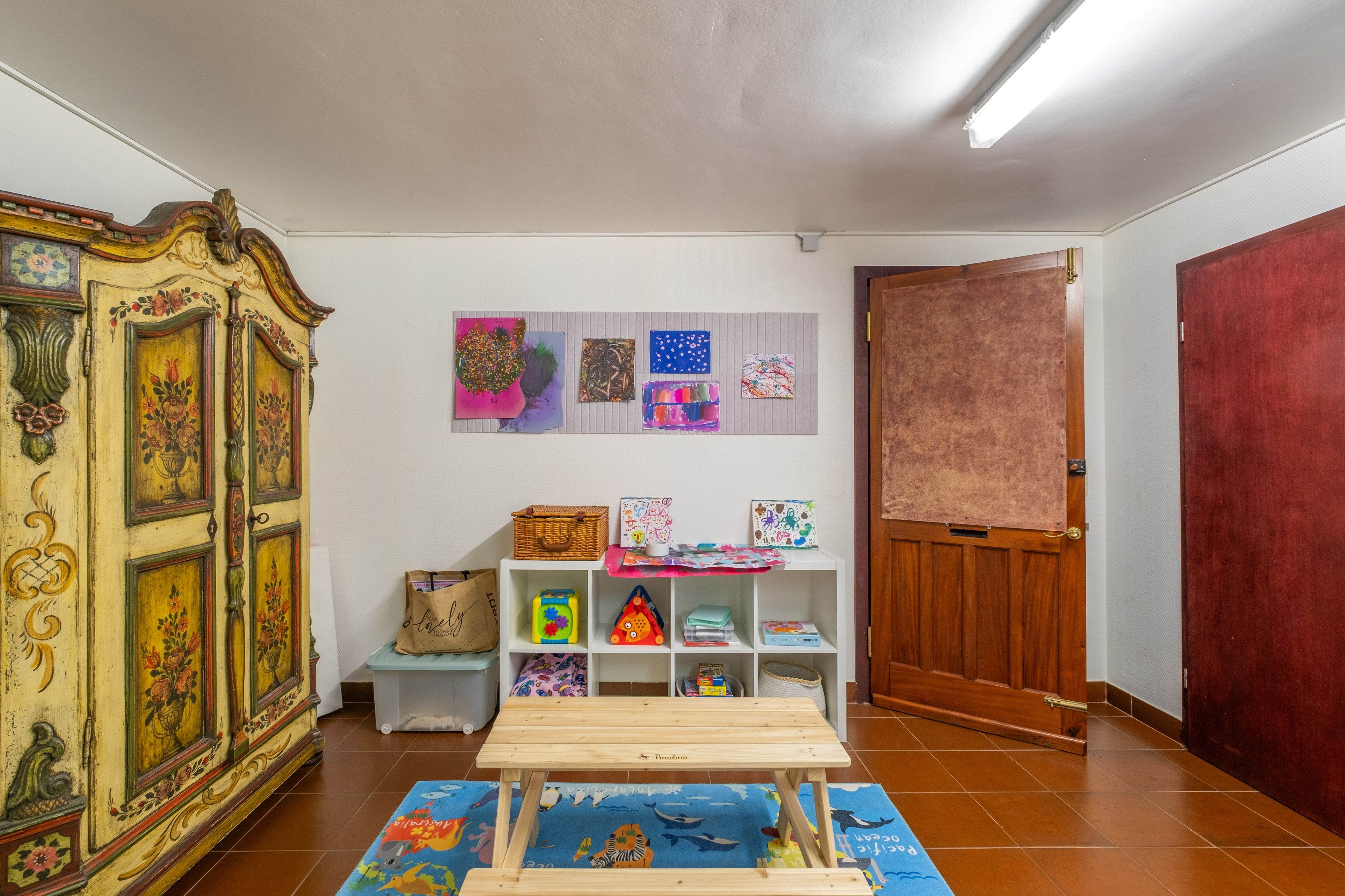 A room with a wooden cabinet, a white shelf with children's artwork, a wooden table, and a red door.