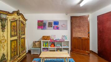 A room with a wooden cabinet, a white shelf with children's artwork, a wooden table, and a red door.