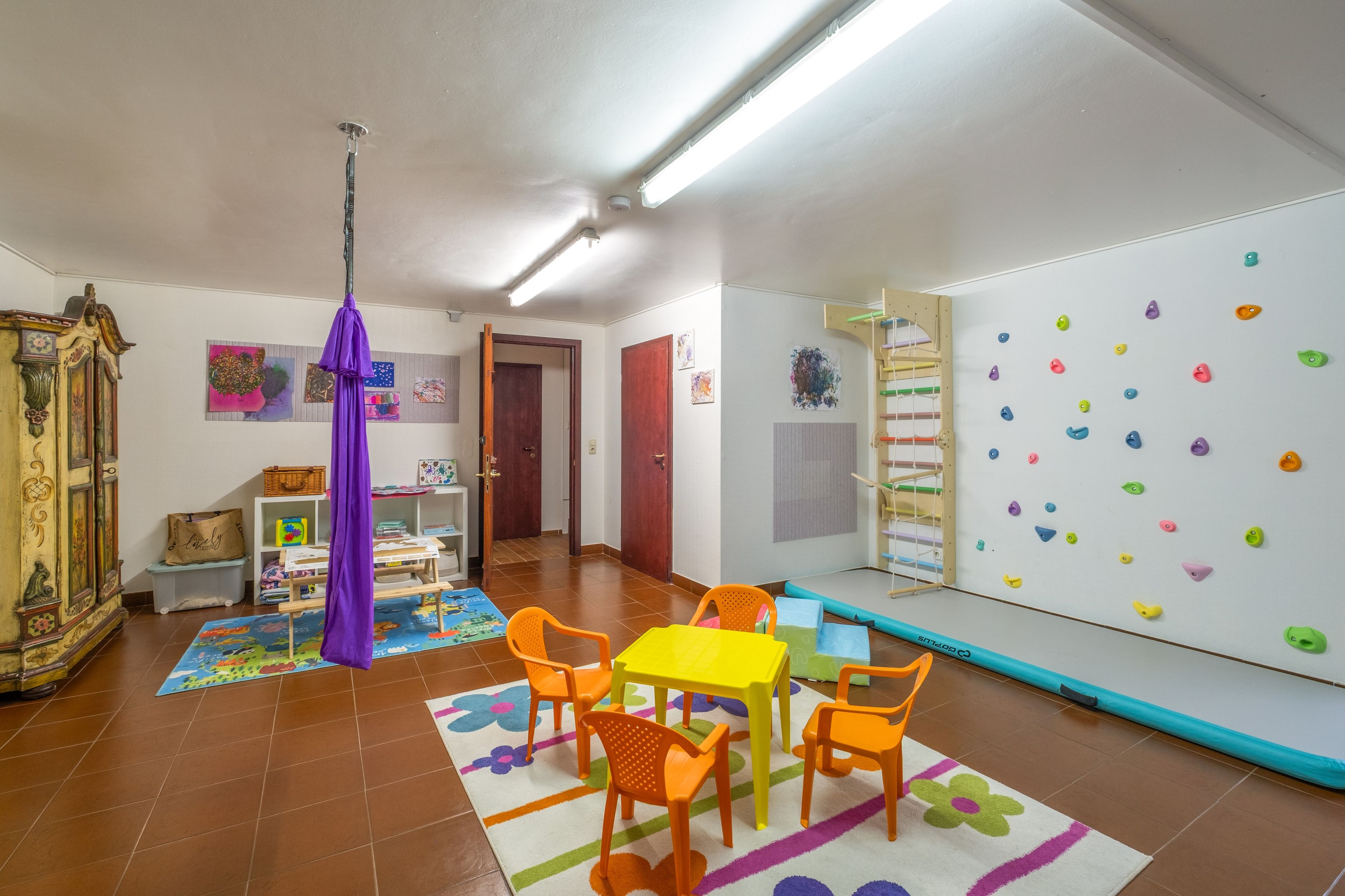 Ein Zimmer mit einer Kletterwand, einem Tisch mit Stühlen, einem violetten hängenden Objekt und einem Bücherregal.