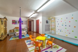 A room with a climbing wall, a table with chairs, a purple hanging object, and a bookshelf.