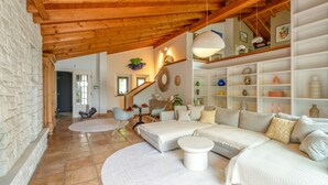 Un ampio soggiorno con soffitto in legno a vista, un divano bianco, un tavolino rotondo e una libreria piena di oggetti decorativi.