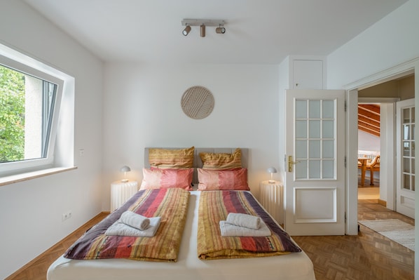 A bedroom with a bed, two bedside lamps, a round wall decoration, and a view of the outdoors through a window.