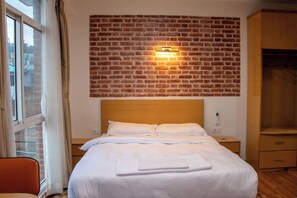 A hotel room with a brick-patterned wall, a bed with white linens, a nightstand, a wooden wardrobe, and a window with curtains.