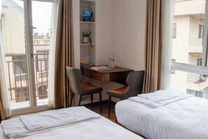A hotel room with two beds, a desk with chairs, a bookshelf, and a window with curtains.