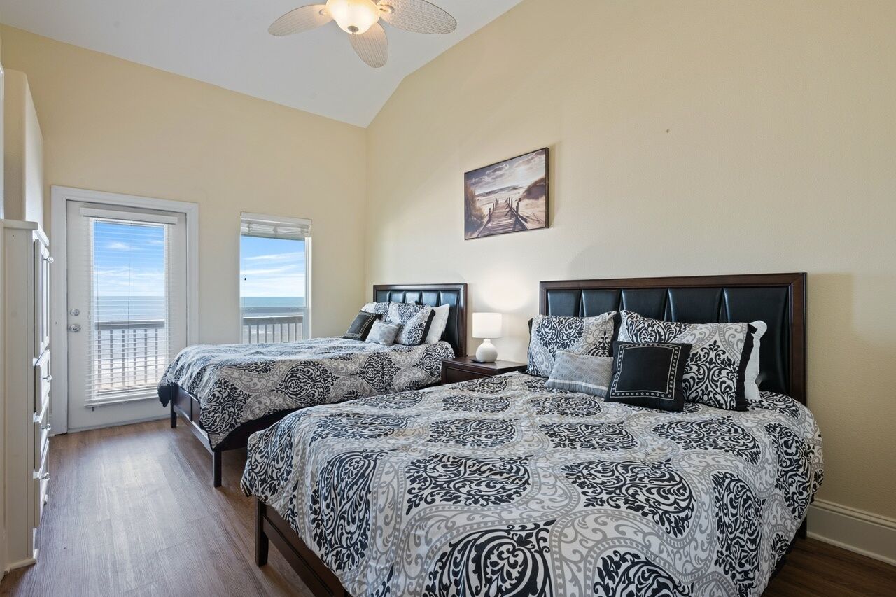 A bedroom with two beds, a ceiling fan, a painting, and a view through a door.