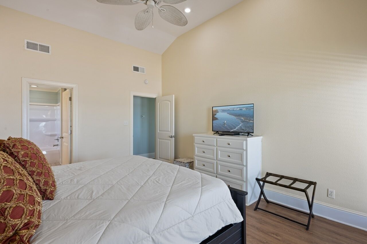 Une chambre à coucher avec un lit, un téléviseur posé sur une commode, un ventilateur de plafond et une porte donnant sur une autre pièce.