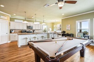 A pool table in a room with wooden flooring, white cabinets, and pendant lighting.