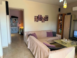 Ein Wohnzimmer mit einer Couch, einem Couchtisch und einem Fernseher. Ein Bücherregal und eine Tür, die in einen anderen Raum führen.
