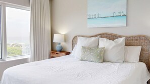A hotel room with a large bed, a nightstand with a lamp, a painting on the wall, and a window with curtains. - Summit 908 – Spacious Gulf-View Retreat at TOPS'L (Miramar Beach)