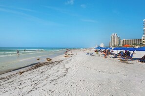 A beach with clear skies, a sandy shore, and a few people. There are umbrellas and lounge chairs, and a building is visible in the background. - Summit 0908A Romantic Gulf-View Studio at TOPS'L Resort (Miramar Beach)