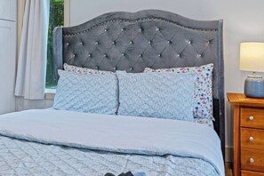 A bed with a tufted headboard, two pillows, a bedside table with a lamp, and a window with curtains. - Tiny Home, HUGE Views, Massive Deck + Fire pit (Blairsville)