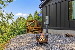 A fire pit in a gravel area with a wooden deck and a building in the background. - Tiny Home, HUGE Views, Massive Deck + Fire pit (Blairsville)