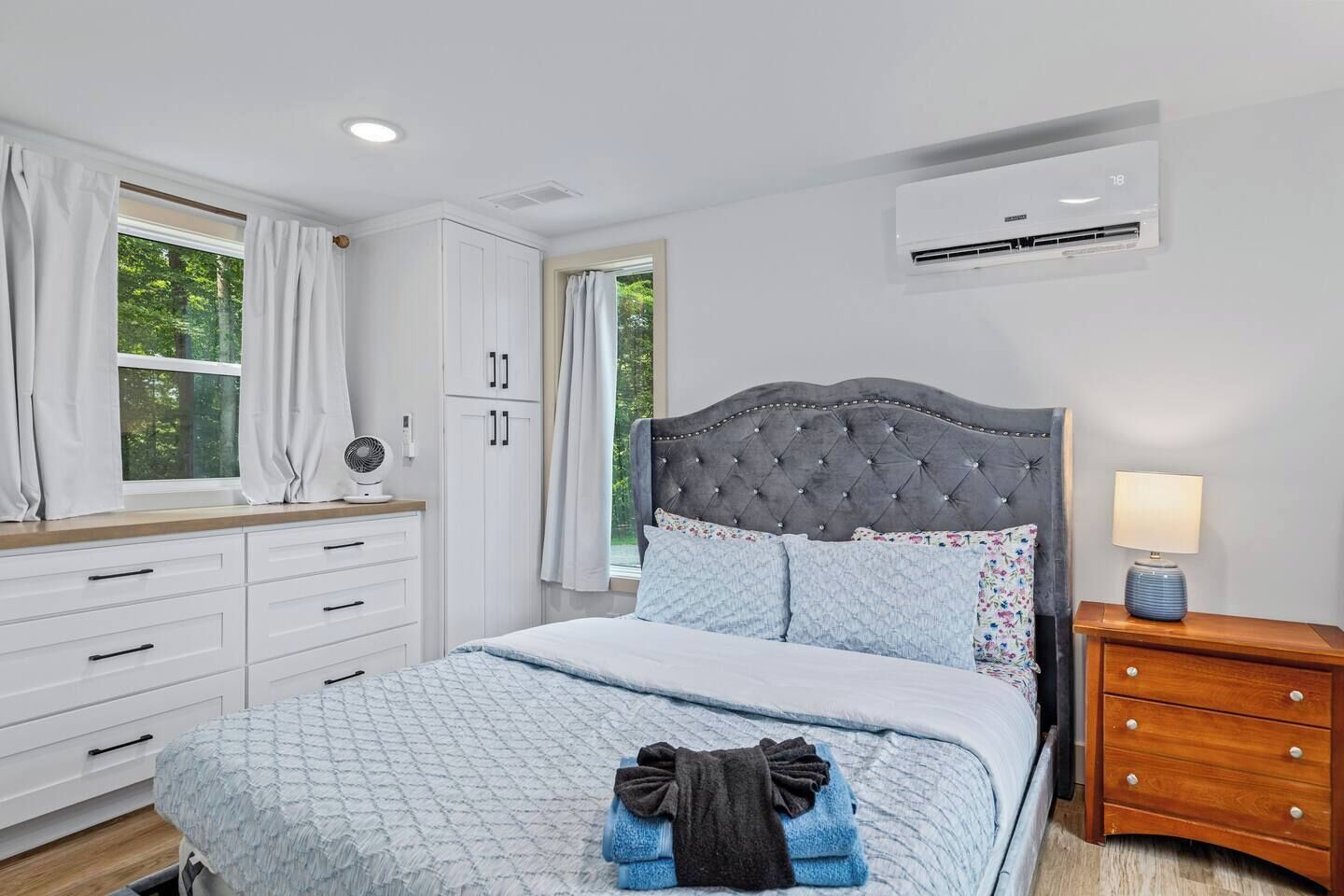 A bedroom with a bed, a bedside table, a nightstand, a window with curtains, a wall-mounted air conditioner, and a ceiling fan.