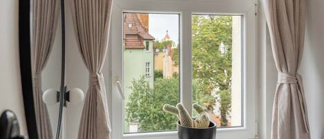 Une fenêtre avec des rideaux, un cactus en pot et une vue sur des immeubles et des arbres.