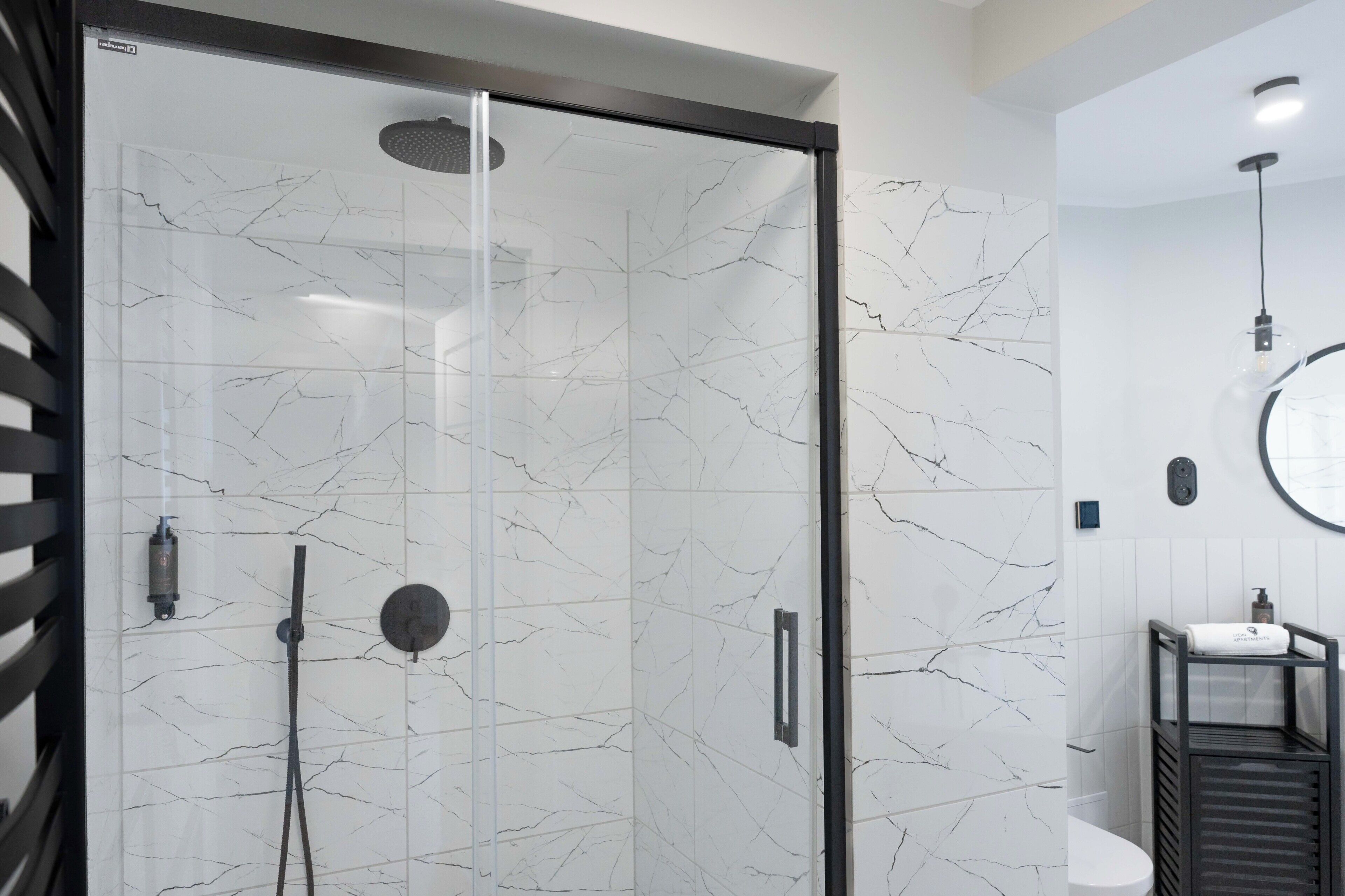 A modern bathroom with a glass-enclosed shower, white marble tiles, a black towel rack, and a round mirror.