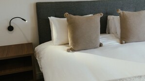 Een opgemaakt bed met witte lakens en twee gestructureerde kussens, een nachtkastje met een lamp en een houten nachtkastje.