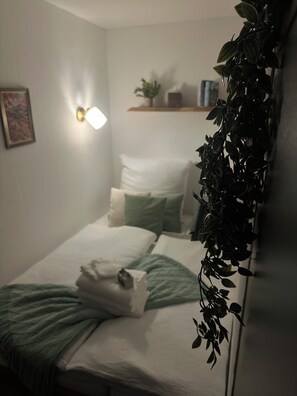 A neatly made bed with pillows and a blanket, a wall-mounted light, and a shelf with a plant. - Ferienwohnung in Kempten – Terrasse & Allgäu (Kempten (Allgäu))