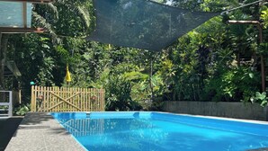 Une piscine rectangulaire, bordée d’une terrasse carrelée, entourée d’une végétation luxuriante et d’une clôture en bois.