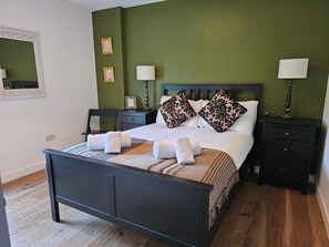 Ein ordentlich bezogenes Bett mit weißen Leinen und dekorativen Kissen, ein Nachttisch mit Lampe sowie ein Sessel in einem Zimmer mit grünen Wänden und Holzdielen.
