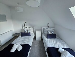 Ein Doppelzimmer mit zwei Betten, jeweils mit Kissen und Decke, einem Nachttisch, einer Lampe und einem Heizkörper.