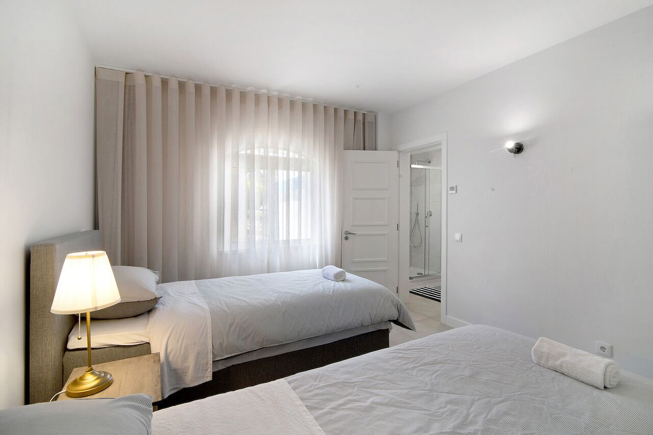 Habitación de hotel con dos camas, una mesita de noche con lámpara, un ventanal con cortinas, una puerta que da a un baño y un aplique en la pared.