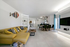 A modern living room with a yellow sofa, a dining area with a table and chairs, and a kitchen in the background. - Beautiful 2-Bedroom Apartment with Private Pool and Garden in Quinta do Lago (Faro)